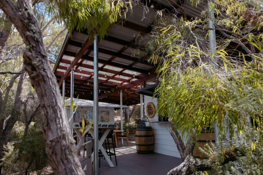 Exterior view of Valle du Venom cellar door nestled among native bushland in Yallingup, featuring a timber and steel pergola with festoon lights and wine barrel decor
