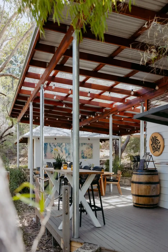Covered outdoor deck at Valle du Venom winery in Yallingup with timber pergola, festoon lighting, long bar-height dining table, and wine barrels alongside native bushland