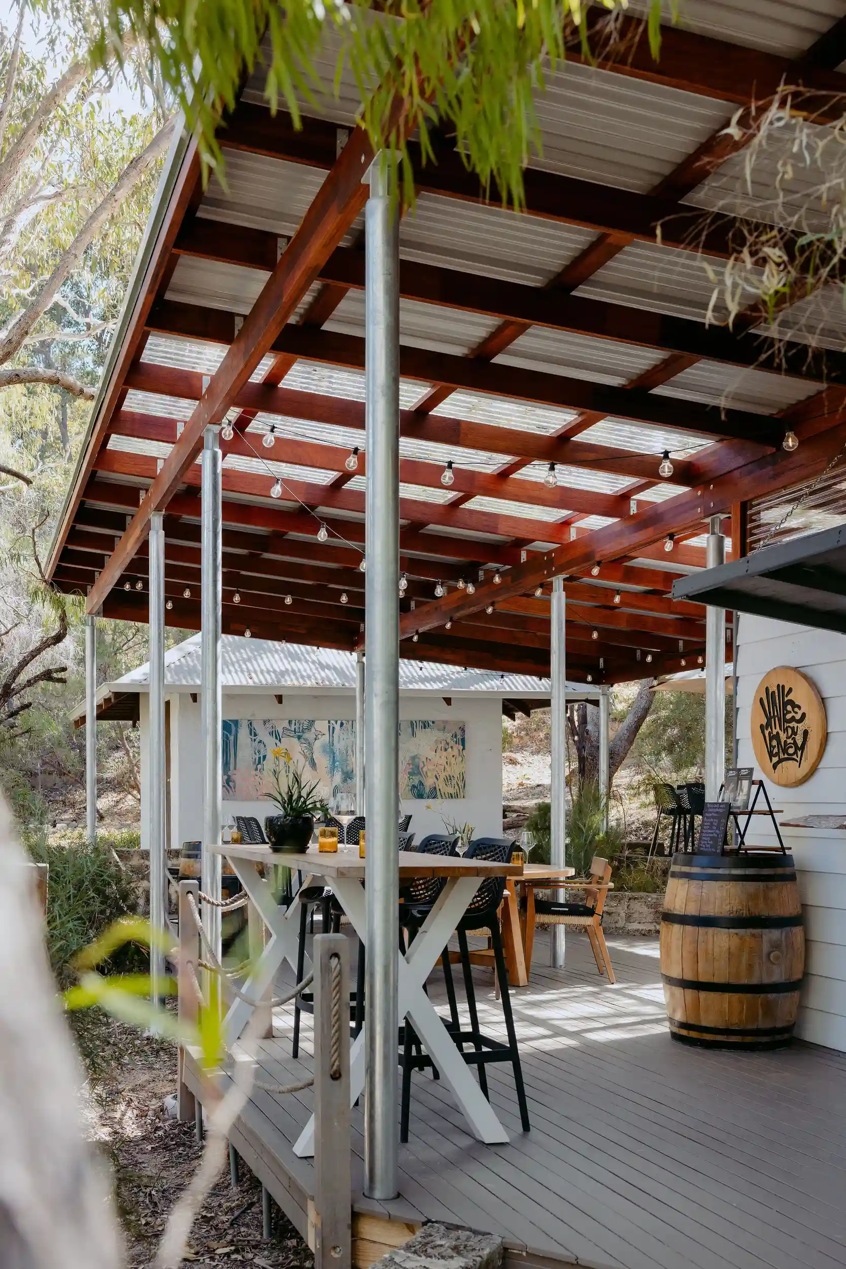 Covered outdoor deck at Valle du Venom winery in Yallingup with timber pergola, festoon lighting, long bar-height dining table, and wine barrels alongside native bushland Covered outdoor deck at Valle du Venom winery in Yallingup with timber pergola, festoon lighting, long bar-height dining table, and wine barrels alongside native bushland