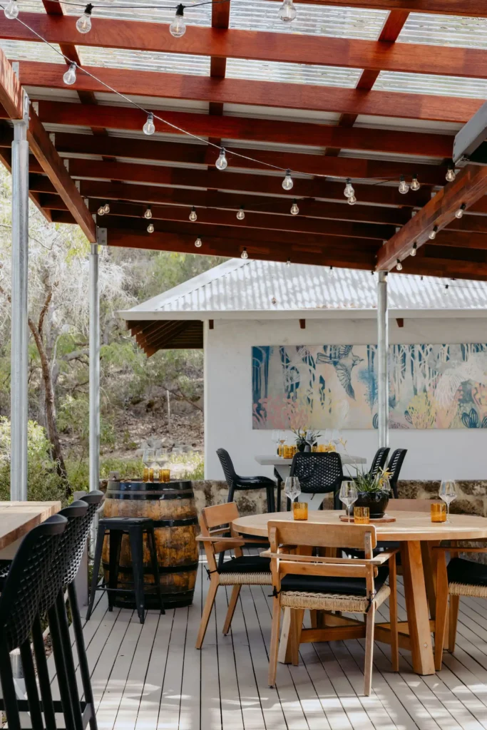 Outdoor dining area at Valle du Venom in Yallingup with round timber table, wine glasses, amber glassware, wine barrel side tables, and large botanical artwork on the back wall