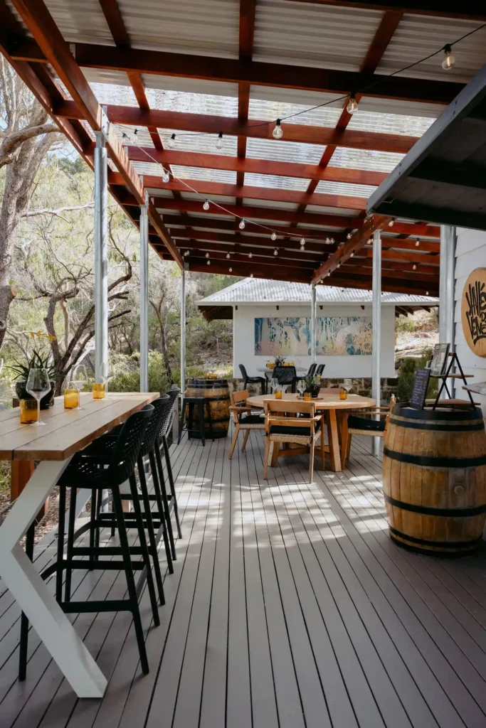 Full view of Valle du Venom winery deck in Yallingup with bar-height seating, round dining tables, wine barrels, festoon lights, and colourful artwork displayed on the back wall