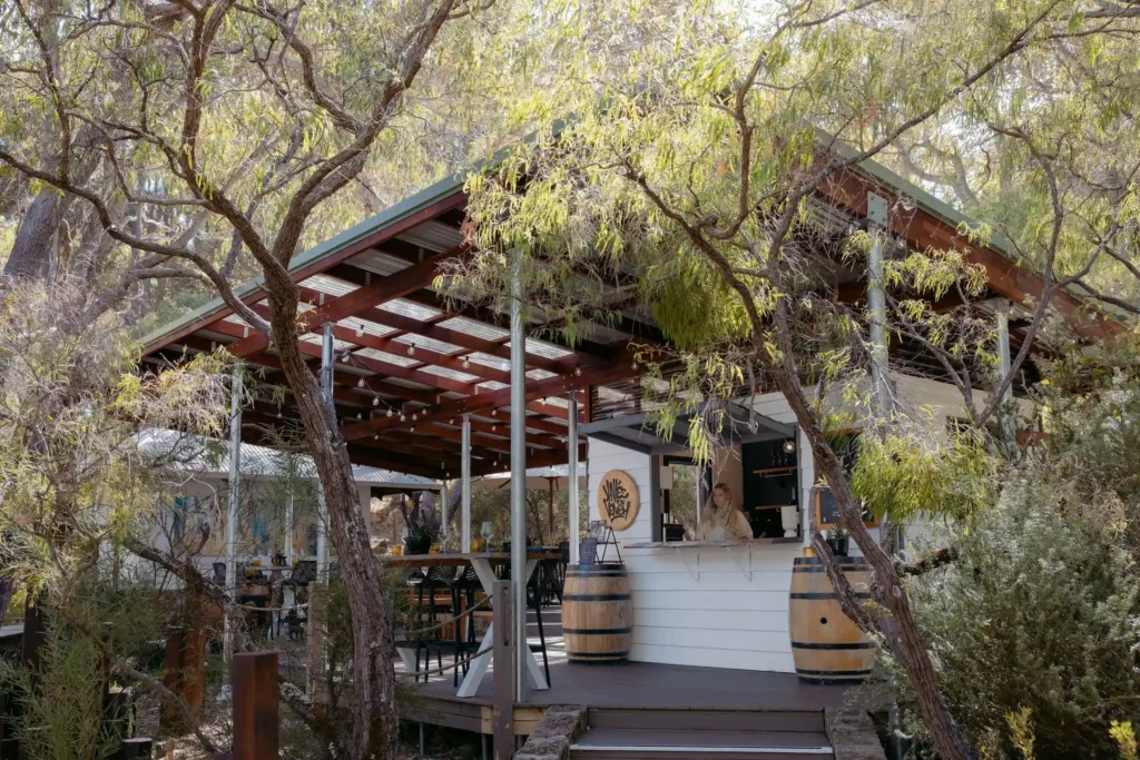 Valle du Venom winery exterior viewed through native Australian trees in Yallingup, with a staff member visible at the cellar door counter and wine barrels on the deck
