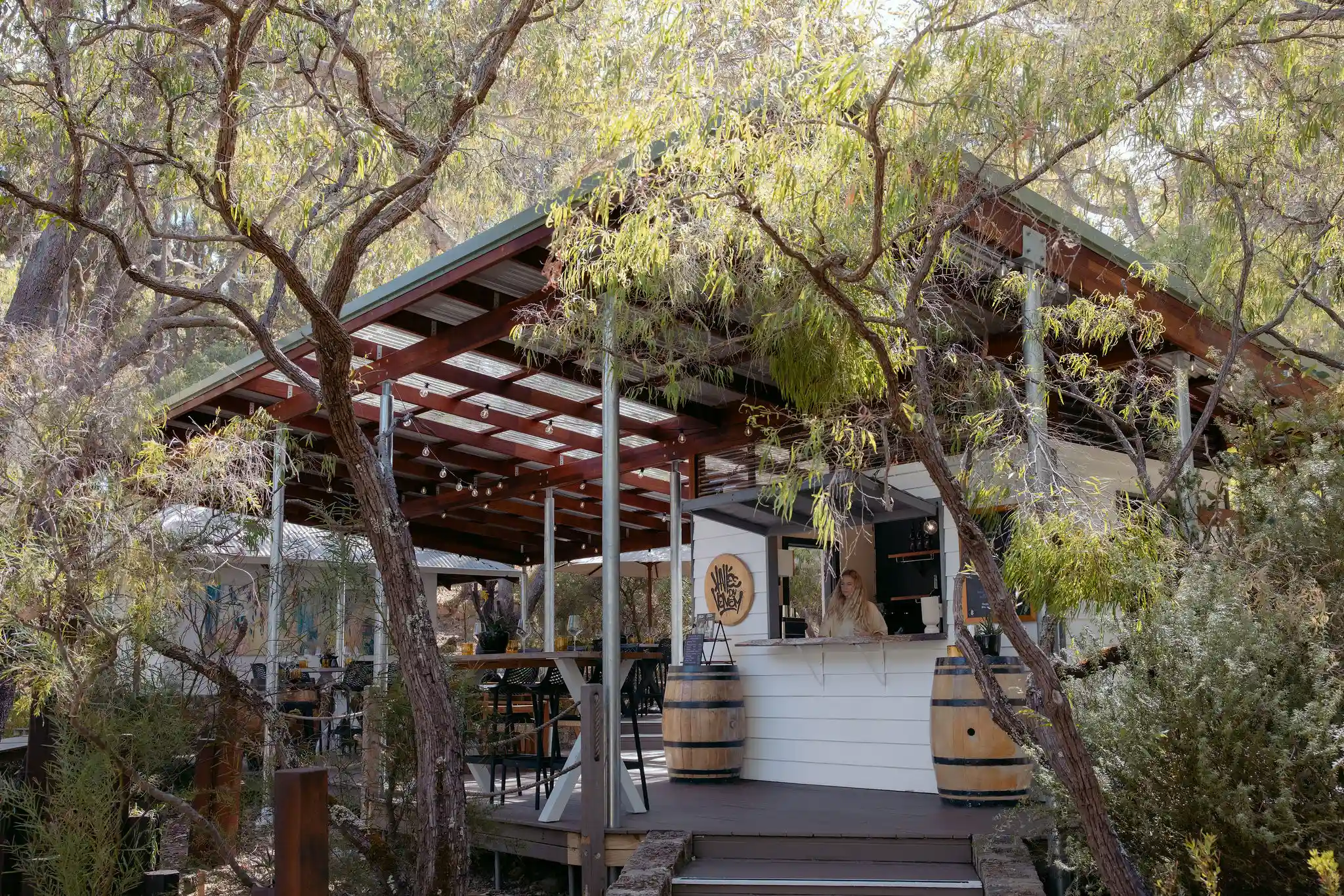 Valle du Venom winery exterior viewed through native Australian trees in Yallingup, with a staff member visible at the cellar door counter and wine barrels on the deck Valle du Venom winery exterior viewed through native Australian trees in Yallingup, with a staff member visible at the cellar door counter and wine barrels on the deck